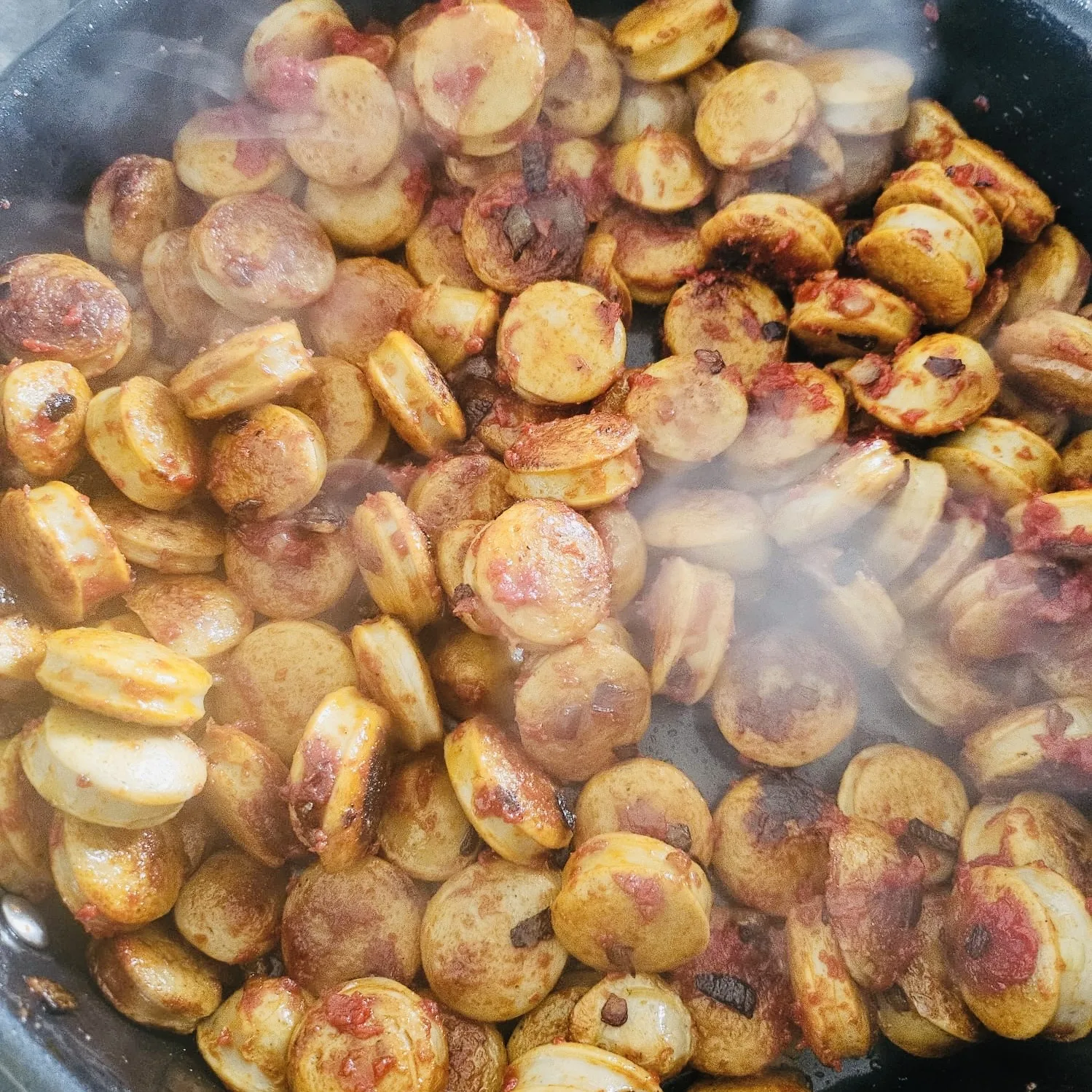 Gebratene Scheiben von Rostbratwurst in einer Pfanne. Die Wurststücke sind kräftig gebräunt und teilweise mit einer würzigen, rötlichen Sauce oder Tomatenpaste überzogen. Aufsteigender Dampf vermittelt den Eindruck eines frisch zubereiteten, herzhaften Pfannengerichts.