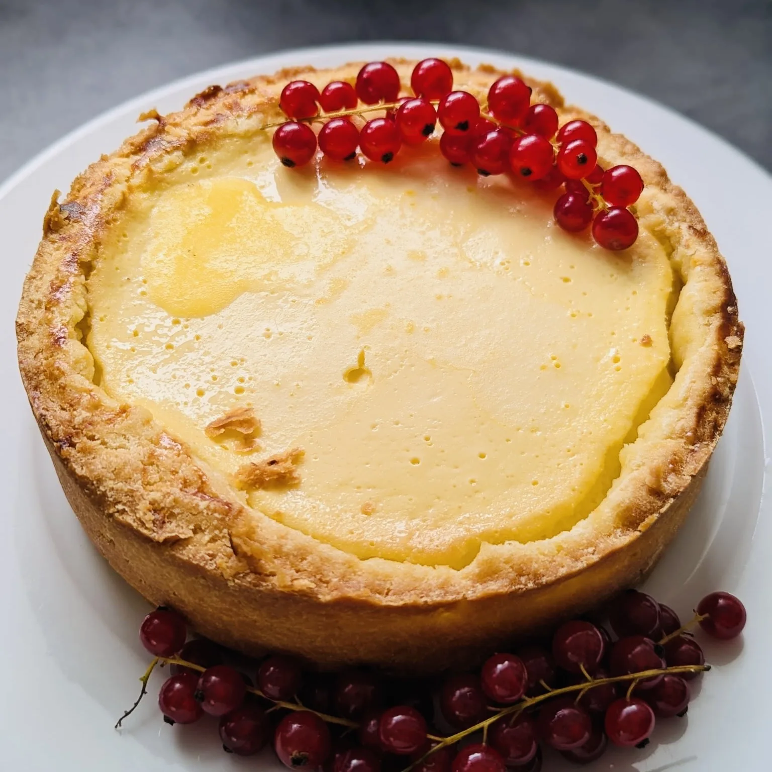 Gebackener Käsekuchen mit hohem, goldbraunem Mürbeteigrand auf einem weißen Teller, garniert mit Rispen roter Johannisbeeren auf der Oberfläche und davor.