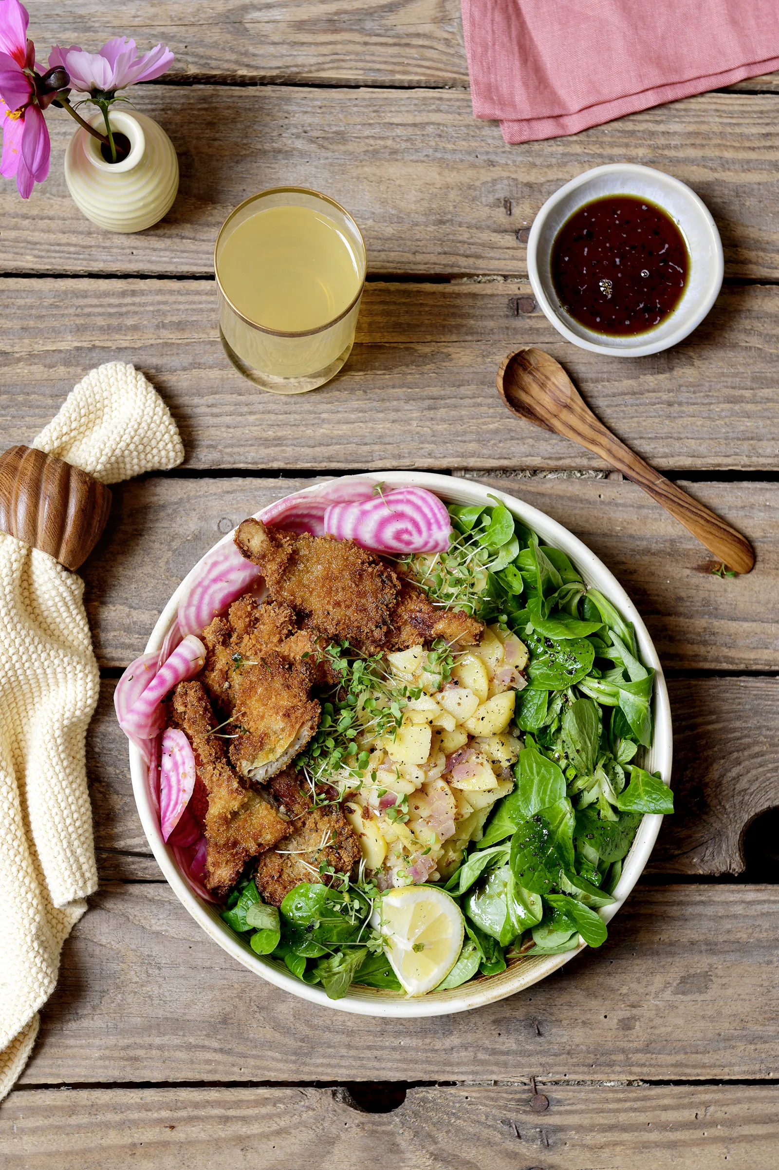Draufsicht auf einen rustikalen Holztisch: In einer hellen Keramikschale liegen goldbraun panierte, knusprig ausgebackene Austernpilze im Backhendl-Style auf frischem Feldsalat, dazu Kartoffelsalat mit roten Zwiebeln und dünne Scheiben pink-weiß geringelter Ringelbete. Obenauf Kresse, am Rand eine Zitronenscheibe. Daneben ein Schälchen mit dunklem Kürbiskernöl-Dressing, ein Holzlöffel, ein Glas mit hellem Getränk, eine kleine Vase mit pinken Schmuckkörbchen, ein rosa Leinentuch und ein cremefarb